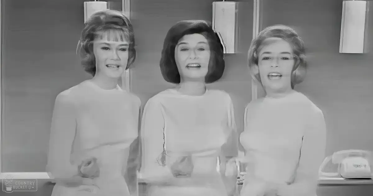 Three young adults in a kitchen setting, wearing light-colored dresses with fitted waists and long sleeves; an older model telephone on the wall suggests it's an older model of the room.