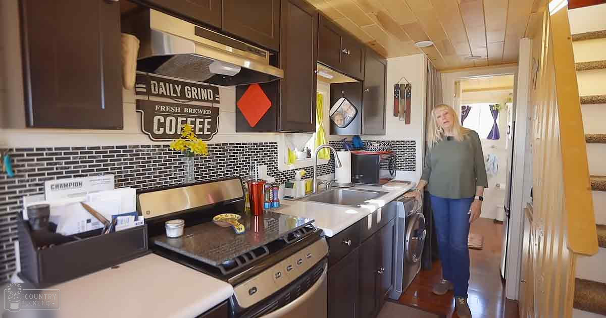 A middle-aged female in a kitchen setting with wooden ceiling panels and a staircase visible. She wears a long-sleeved, loose-fitting green top and blue jeans.