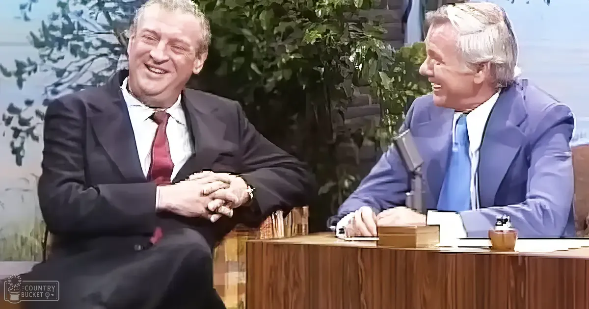 Two adults in a talk show setting with greenery backdrop, bright lighting, and professional attire from past decades.