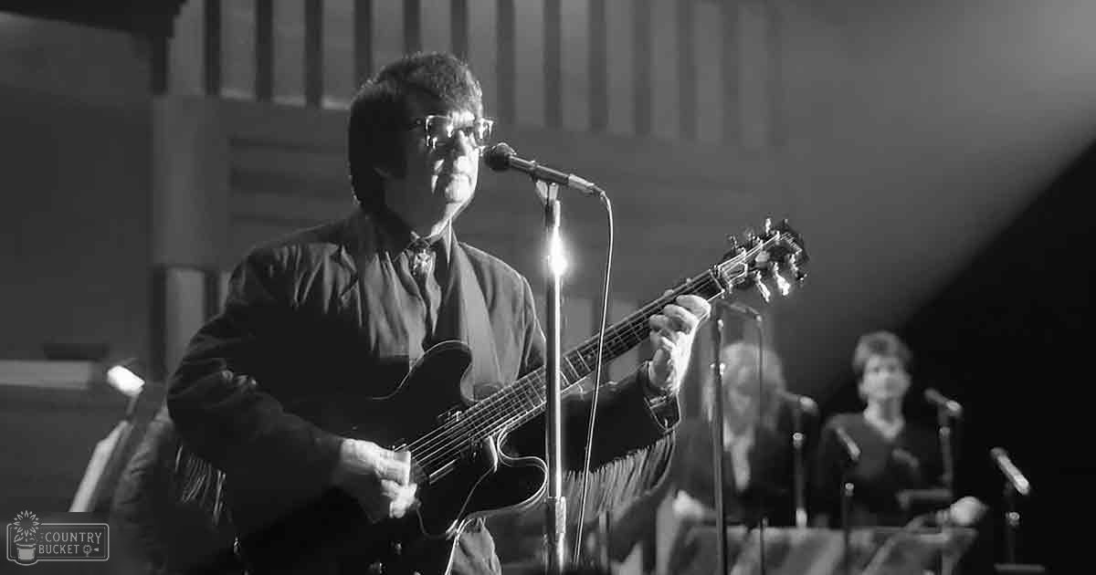 Middle-aged man playing guitar in an indoor concert setting with a middle-aged woman in the background, wearing dark attire and microphones visible on stands.