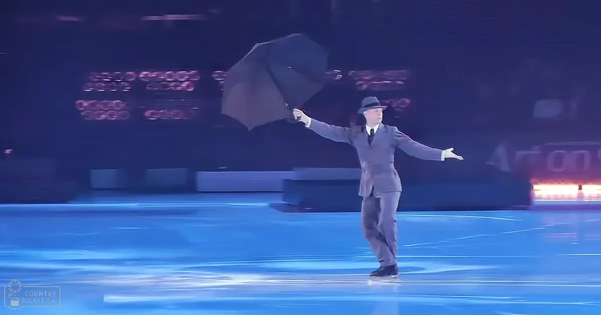 An older adult male performer in a dark gray suit and fedora holding a black umbrella on an indoor ice rink with stage lighting.