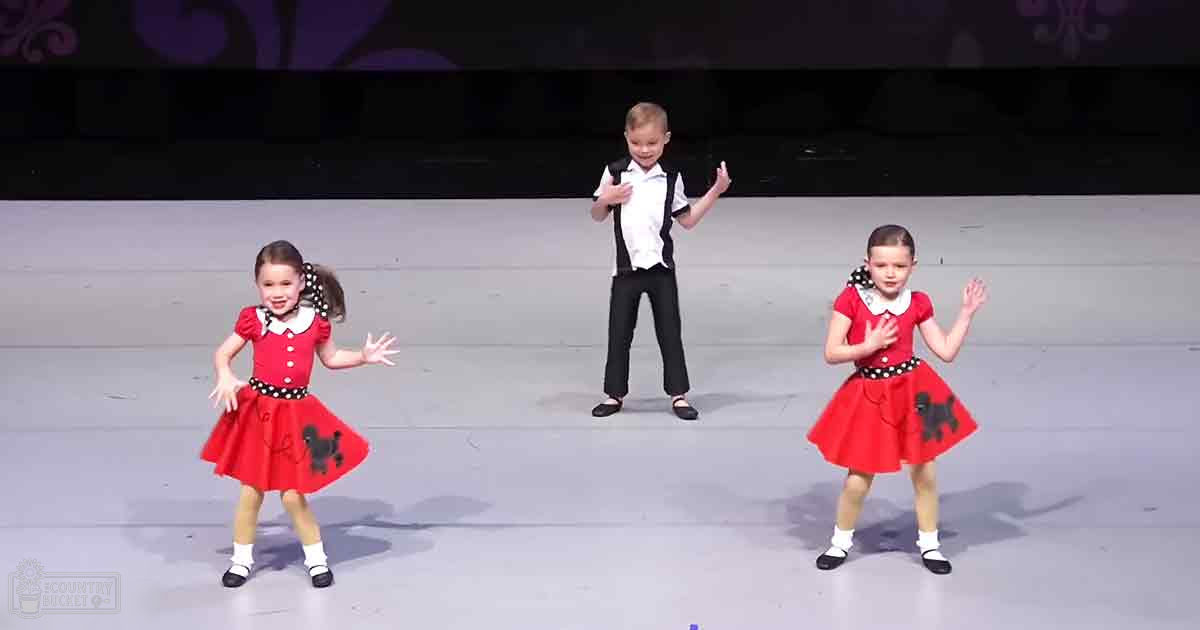 Children in a toddler/child age band perform on stage at an indoor venue with bright lighting, likely during daytime or under artificial lights.