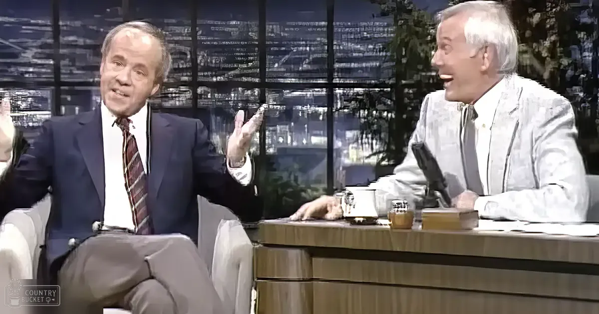 A talk show host in a studio setting, wearing a dark blue suit jacket with red plaid tie, alongside an older adult male in a light patterned suit, both seated at a desk with a microphone and urban nightscape visible through the windows.