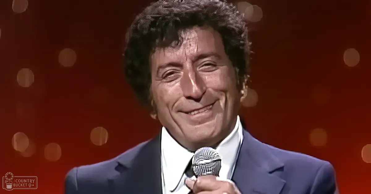 A middle-aged adult male performer wearing a dark blue suit jacket with a white shirt and black tie, standing on stage or in a studio setting, holding a microphone under warm-toned bokeh lighting.