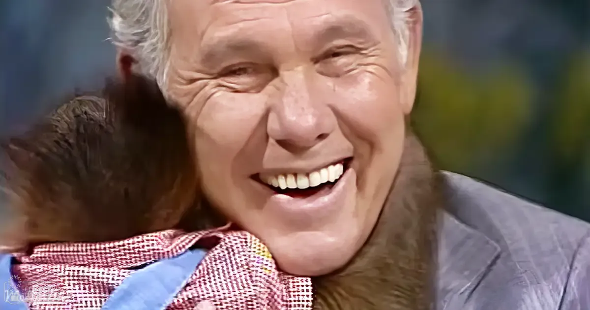 An older adult male wearing a light blue, textured square-patterned shirt indoors with a toddler and a dark brown cat nearby under artificial lighting.