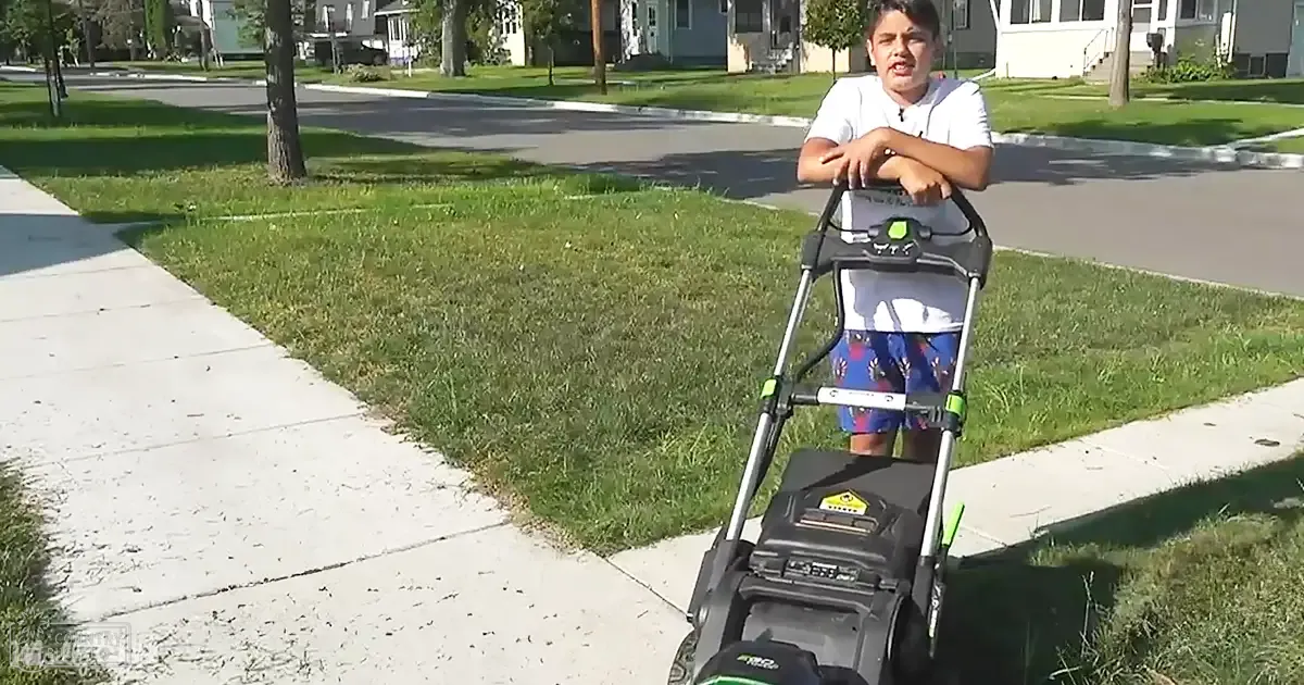 A young boy mowing grass in a suburban neighborhood under clear, sunny skies on what appears to be summer or early fall.