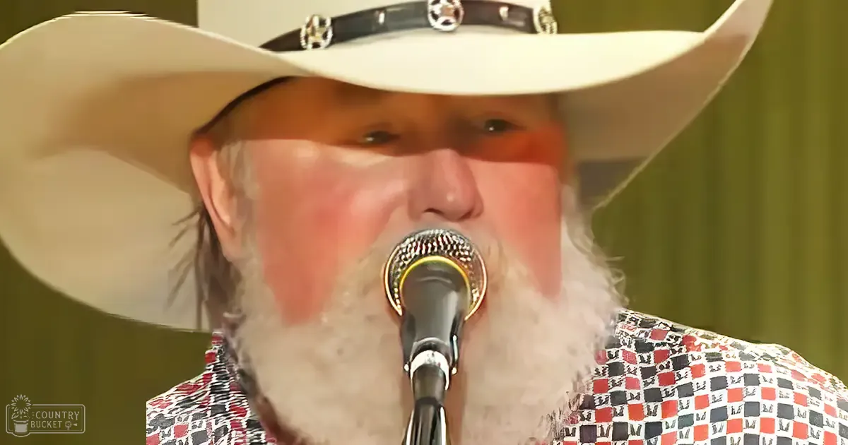 A middle-aged adult male wearing a relaxed fit, collared, long-sleeved cowboy shirt with a red, black, and white geometric checkered pattern in what appears to be an indoor performance or recording venue.