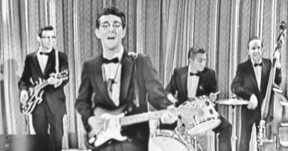 A young adult male in a black suit jacket, white shirt, and black bow tie plays an electric guitar on stage with other band members in similar attire.