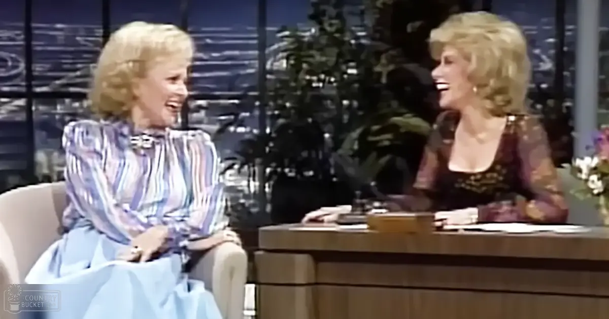 Talk show studio setting with an older adult female in a striped dress with ruffled sleeves, paired with another older adult female wearing a dark-colored lace dress with intricate patterns at the neckline and shoulders. The backdrop suggests it's evening or night time.
