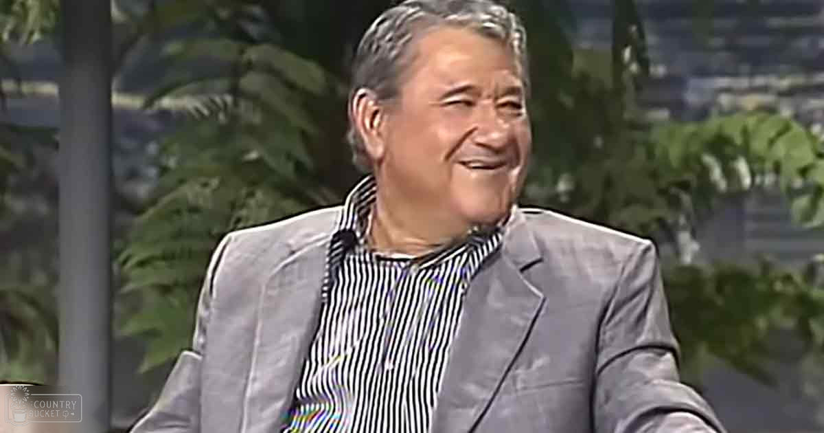 An older adult male in a formal gray suit and black-and-white striped shirt, seated on a couch in an indoor studio setting with greenery in the background.