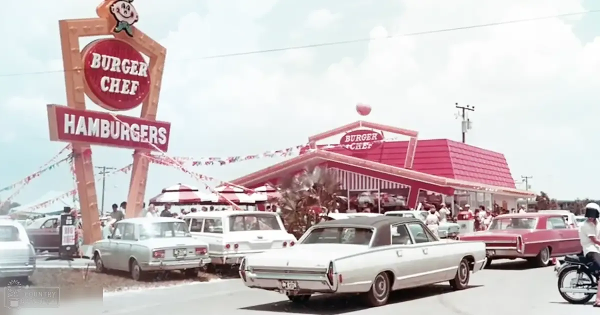 A Burger Chef restaurant in an outdoor setting with palm trees and 1960s-era cars parked outside, partly cloudy sky suggesting midday or early afternoon.