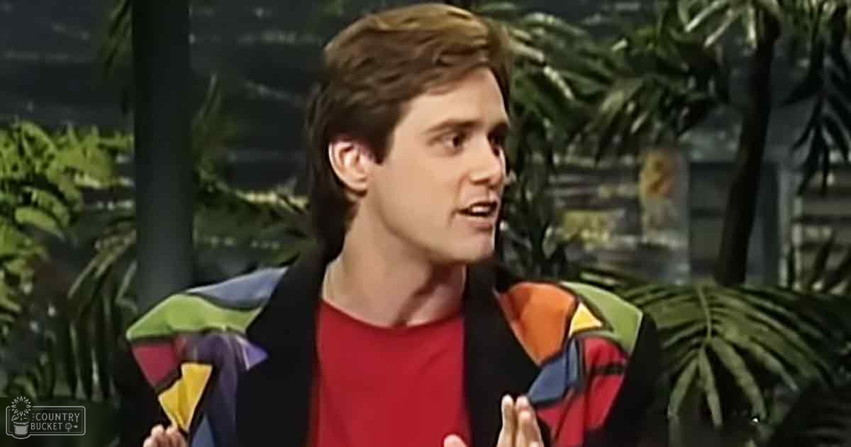 A young adult male in a relaxed fit, colorful jacket over a red shirt with geometric patterns stands indoors amidst lush greenery under natural daylight.