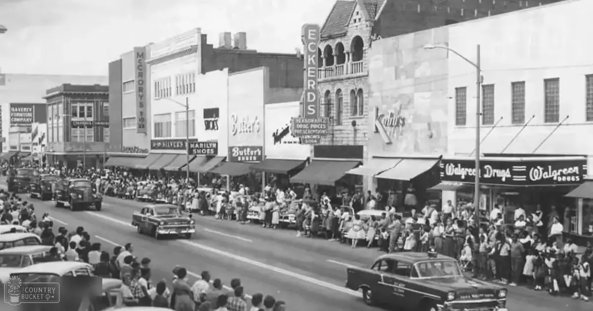 A parade scene in an urban setting with mid-20th century architecture, with various clothing styles from infants to older adults and vintage vehicles like a Chevrolet Bel Air and Ford Police Interceptor.
