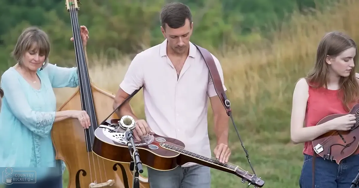 Musicians performing outdoors in a natural setting with grass and trees, late afternoon light.
