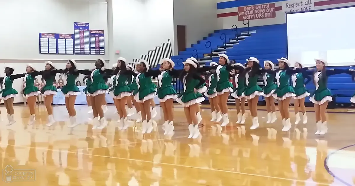 A group of young adult female cheerleaders in green, shiny synthetic uniforms with ruffled skirts and white cowboy boots standing indoors in a gymnasium setting.