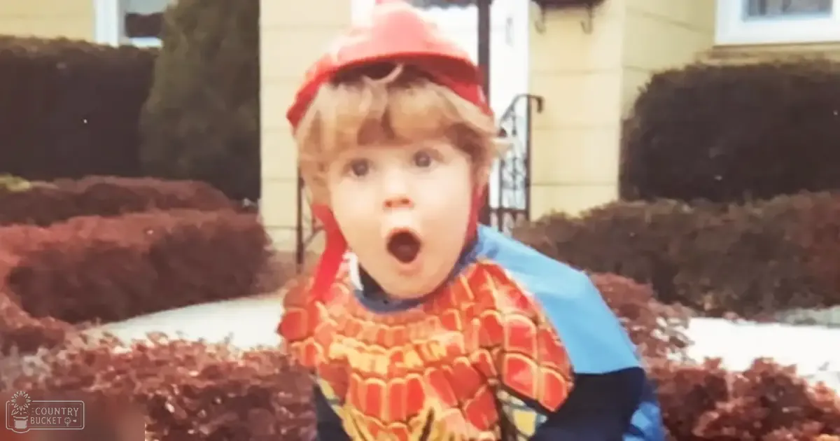 A toddler in a Spider-Man costume stands outdoors near a house with bushes and a paved surface, suggesting spring or fall.