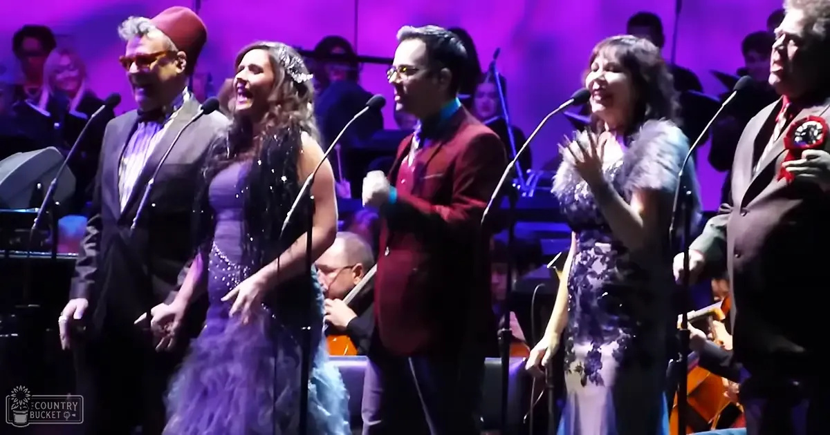 An indoor concert hall performance scene with musicians and singers wearing formal attire, including a red fez hat and sequined dress, under stage lighting during what appears to be a live music event.