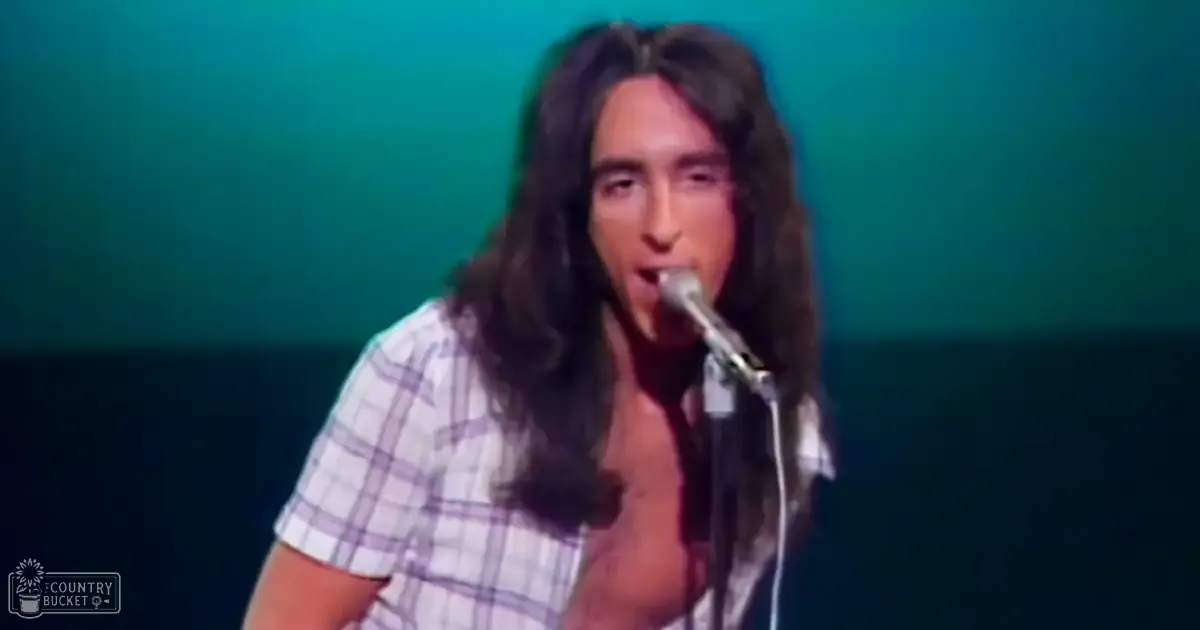 A young adult male singer performs on stage in front of a microphone against a solid teal backdrop under stage lighting, wearing a white and blue plaid short-sleeved shirt from the 1970s.