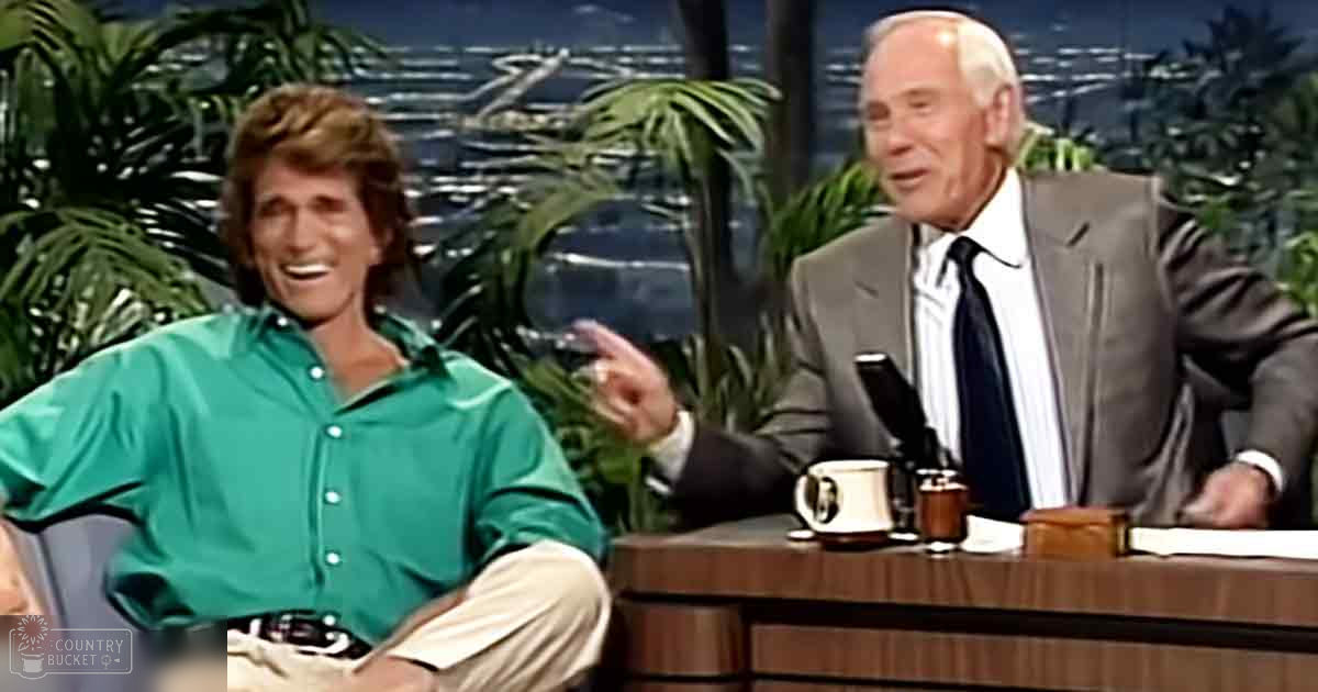 Two adults, one in a green button-up shirt and beige pants, the other in a gray suit jacket over a white dress shirt with a black tie, seated at a desk against a backdrop of tropical plants and a nighttime cityscape, representing an indoor talk show set.