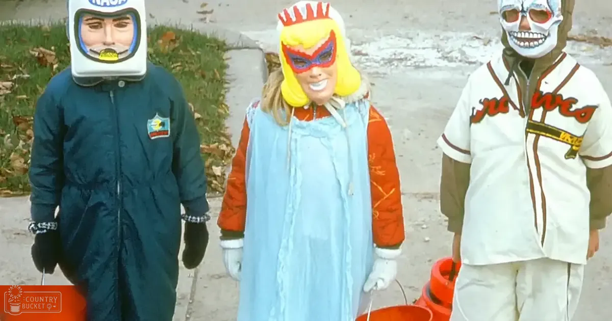 Children in Halloween costumes, including a dark green jumpsuit with patches and helmet mask resembling an astronaut, light blue dress with orange sleeves and white gloves, and white baseball-style jacket with brown stripes and skull-like designs. Each holds an orange bucket on a residential street during autumn.