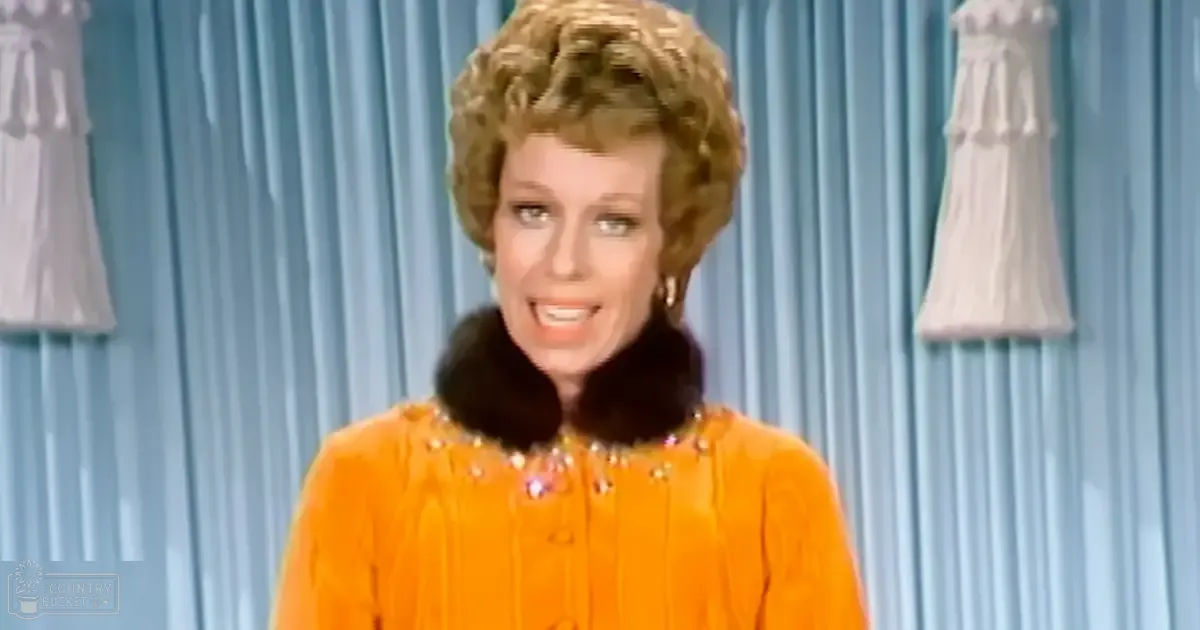 A middle-aged female in an indoor setting, wearing a bright orange dress with fur collar and embellishments. Two white dresses hang against blue curtains.