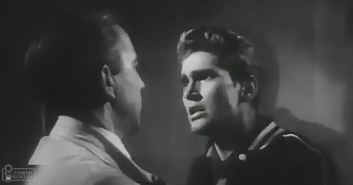 A mid-20th century film scene with two adult male actors dressed casually against a plain backdrop in an indoor studio environment.
