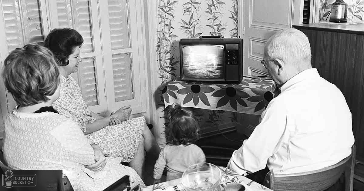 A family of four watching television in a mid-20th-century living room with wooden shutters and leafy wallpaper.