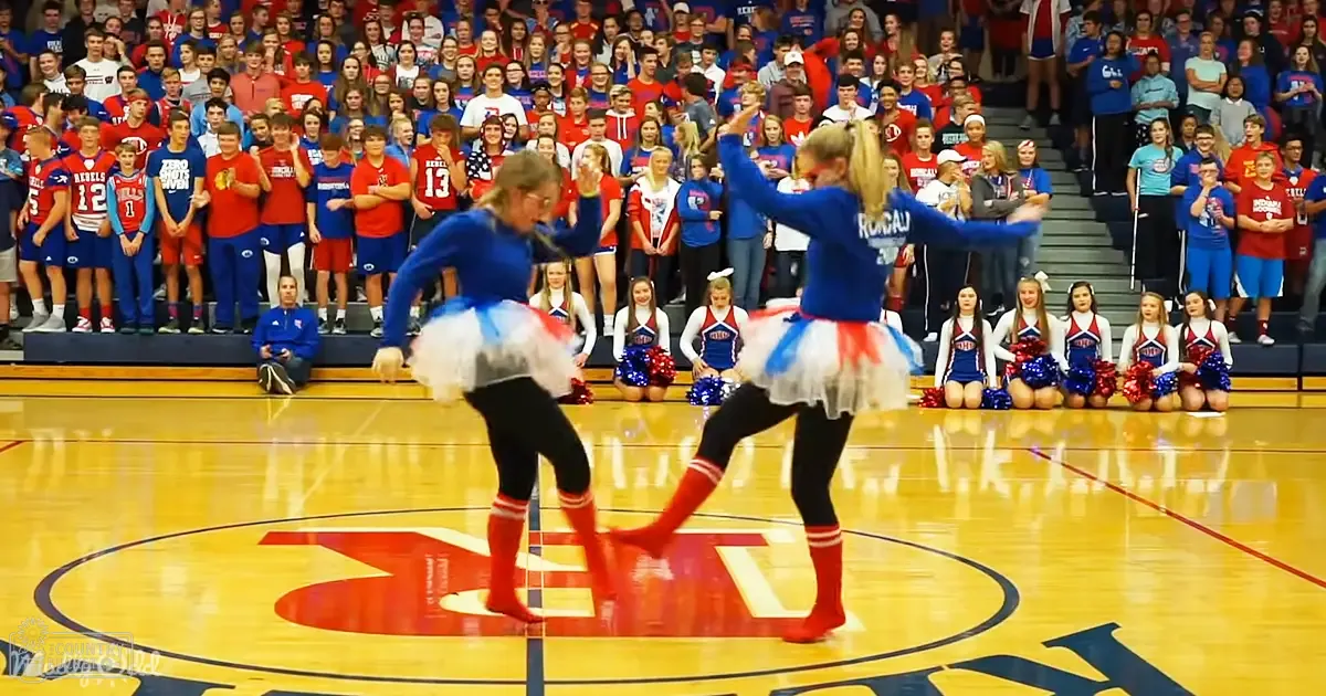 A dance performance at an indoor sports arena with children and adults in school uniforms, highlighting a vibrant red, blue, and white color scheme.