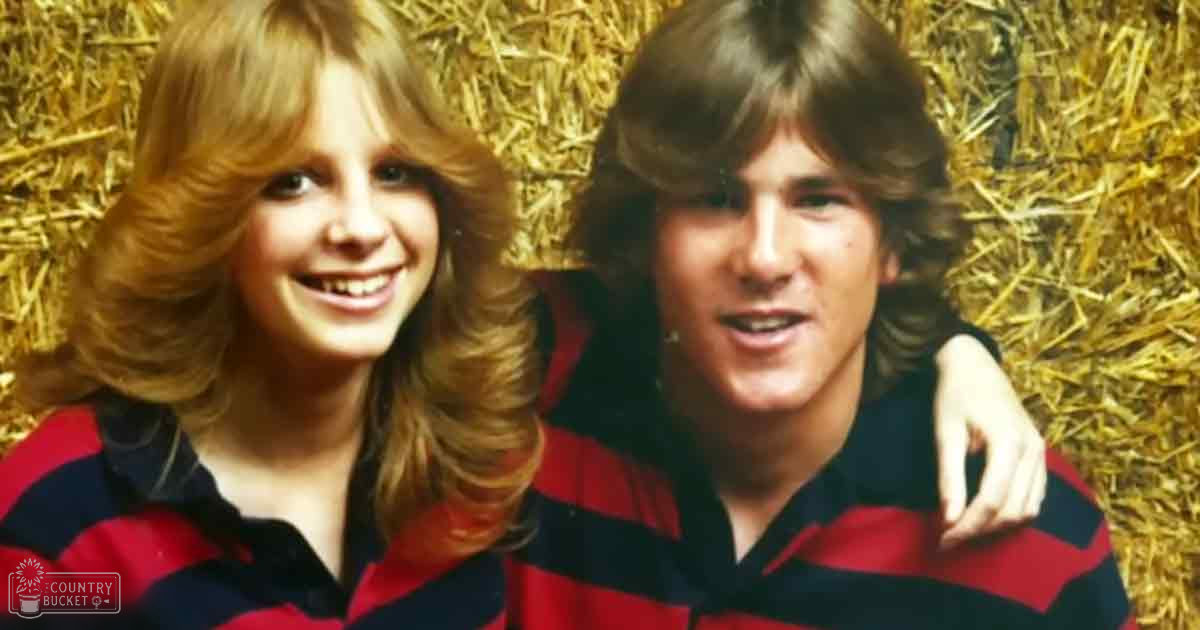 A young couple wearing red and navy blue striped polo shirts, standing indoors with hay bales as background, suggesting a barn or stable setting under natural light.