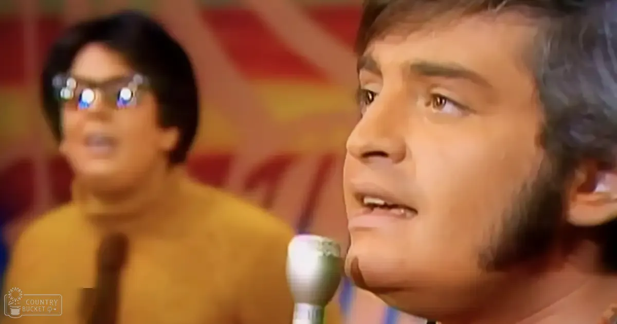 A young adult male singer wearing a dark, 1970s-style shirt with long sleeves and high collar performs into a microphone at an event or performance. Another individual in the background wears glasses and a yellowish-brown turtleneck sweater.