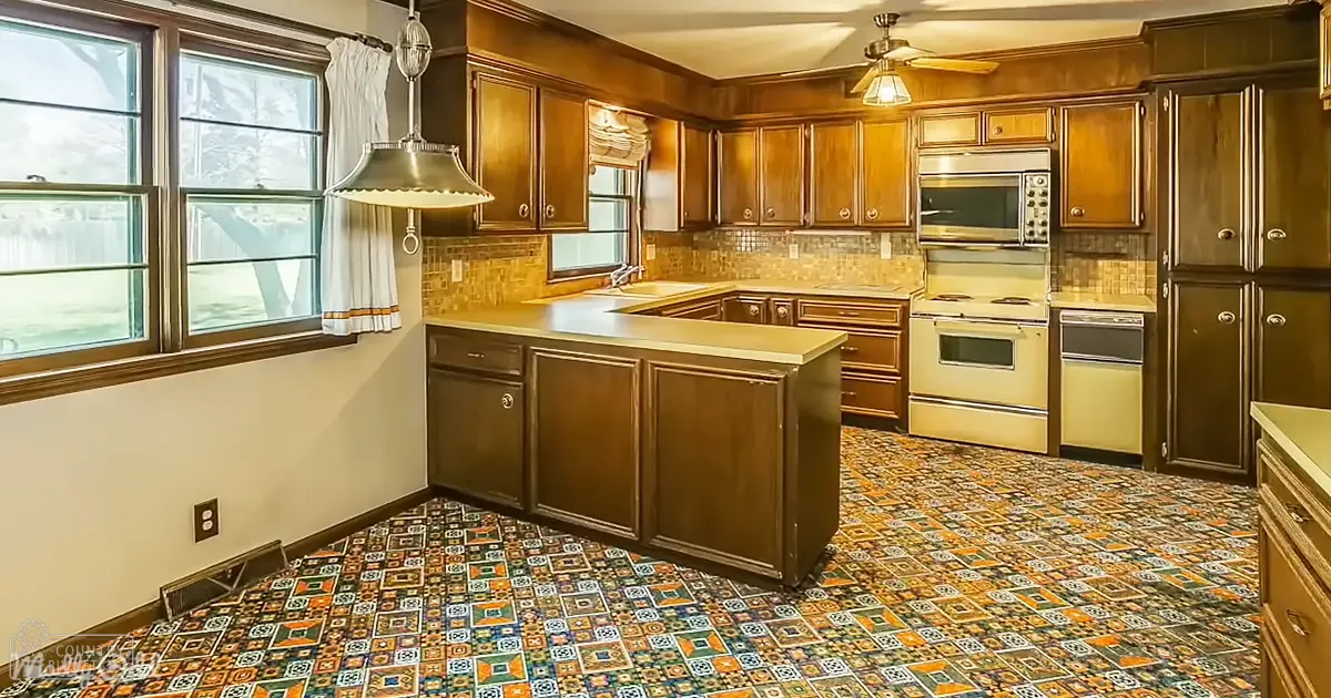 A residential kitchen with wooden cabinets, a central island counter, a range hood, and patterned tile flooring. Natural light streams in through windows with curtains.