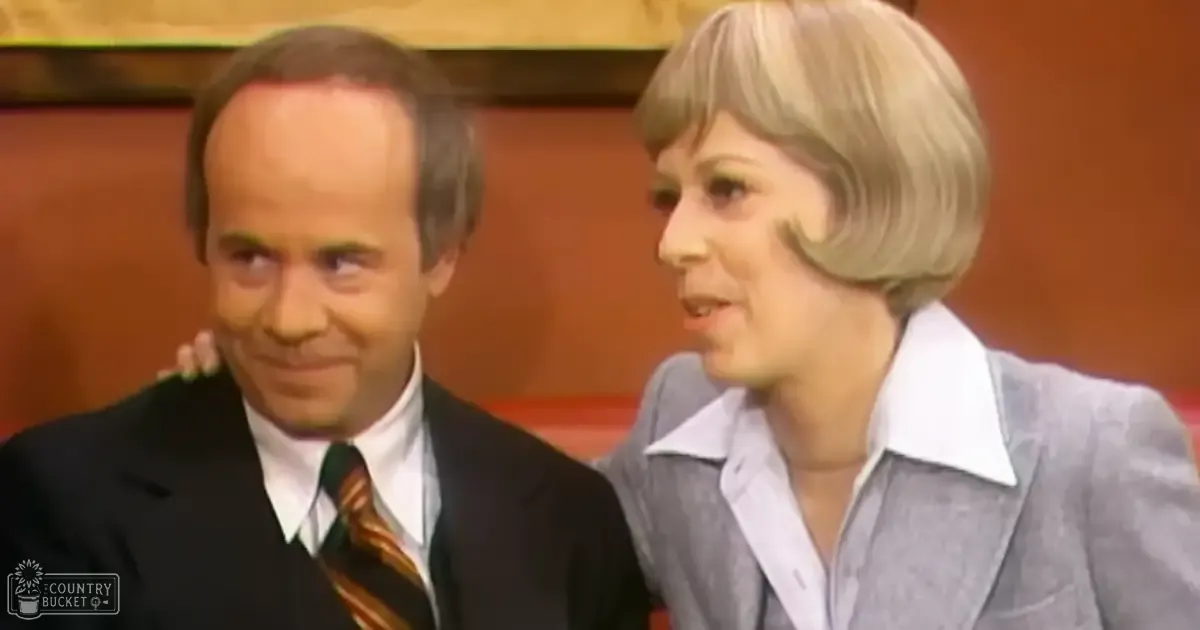 A middle-aged man in a dark suit with a striped tie stands alongside an older adult woman wearing a light gray blazer, both positioned indoors, possibly on a stage or set.