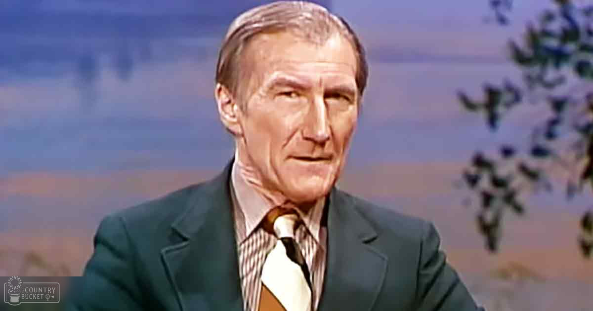 An older adult male wearing a dark green, tailored suit jacket with a light-colored, striped shirt in what appears to be a studio setting designed to mimic an outdoor scene.