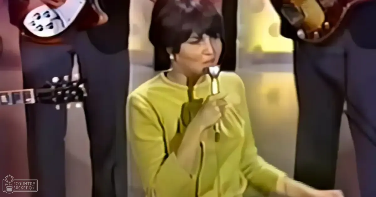 A young adult female wearing a bright yellow vintage-style long-sleeved top with a high collar and bow, holding a microphone indoors under bright artificial lighting.
