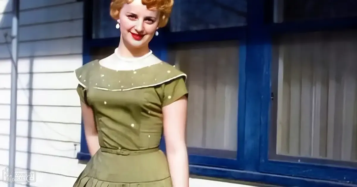A young adult female wearing a mid-20th-century, olive-green polka-dot dress with cap sleeves stands outside in front of a house with white siding and blue-framed windows. The scene is brightly lit, suggesting spring or summer.