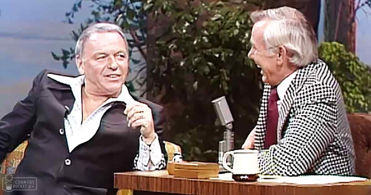 A talk show set with two middle-aged to older adult men in formal attire against a backdrop of trees and possibly water, suggesting an outdoor scene painted onto the set. The lighting is natural daylight.