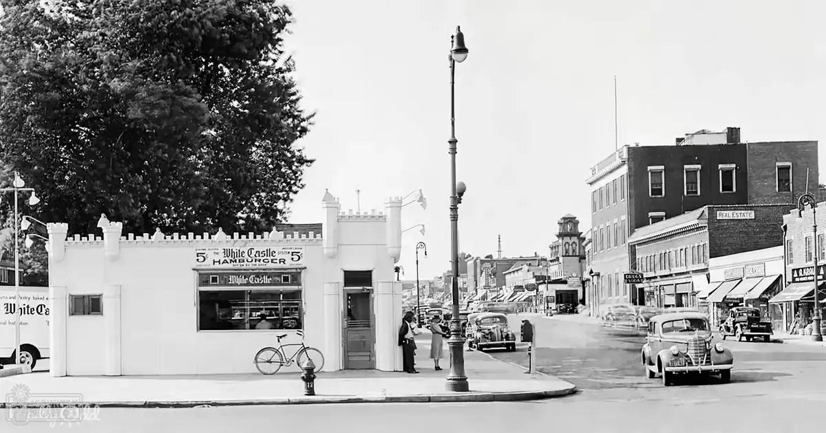 A nostalgic urban street scene with vintage cars, a White Castle hamburger sign, and ornate streetlights in a mid-20th century small town or city setting.