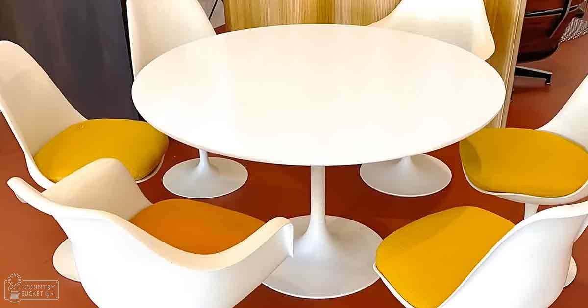 A modern dining set with a white pedestal table, six white plastic-framed chairs with yellow cushions, against wooden paneling in an indoor setting.