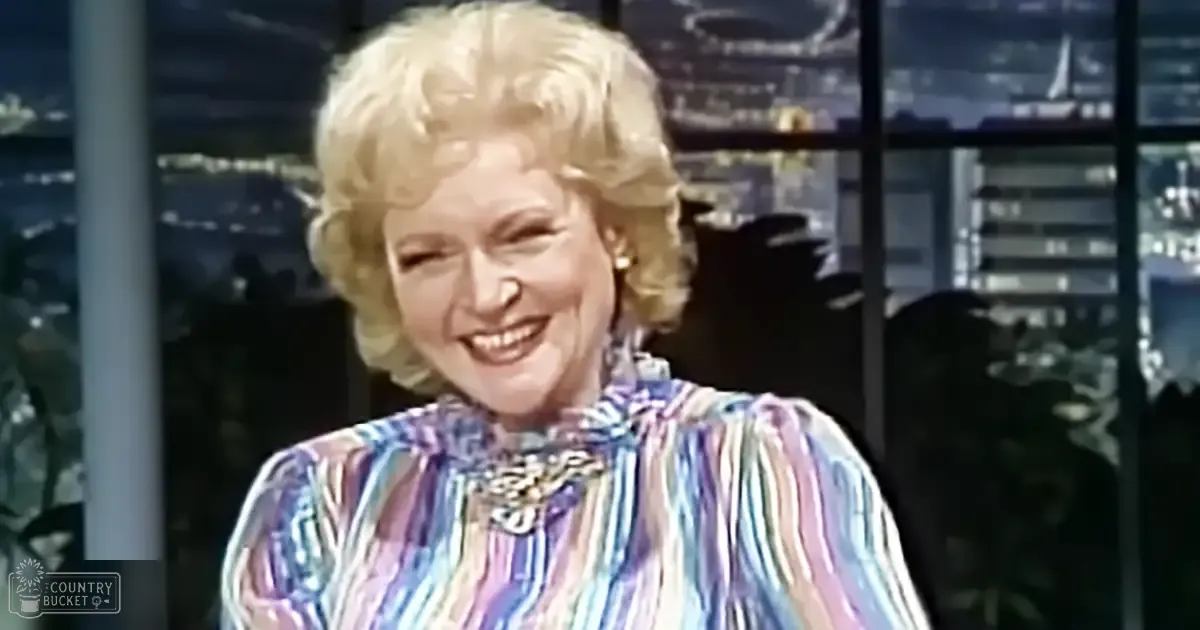 Older adult female wearing a colorful, striped blouse with ruffled details and gold earrings in a studio setting with large windows showing an urban night scene outside.
