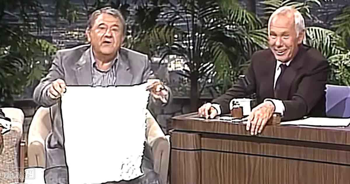 A middle-aged man in a gray suit jacket holds a white cloth or paper while an older adult male guest, dressed in a black suit jacket and white shirt with tie, sits at a wooden desk in a television studio designed to mimic an outdoor environment.