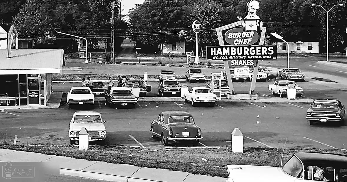 Burger Chef drive-in restaurant with vintage cars and 1960s attire adults in an urban parking lot on a sunny day.