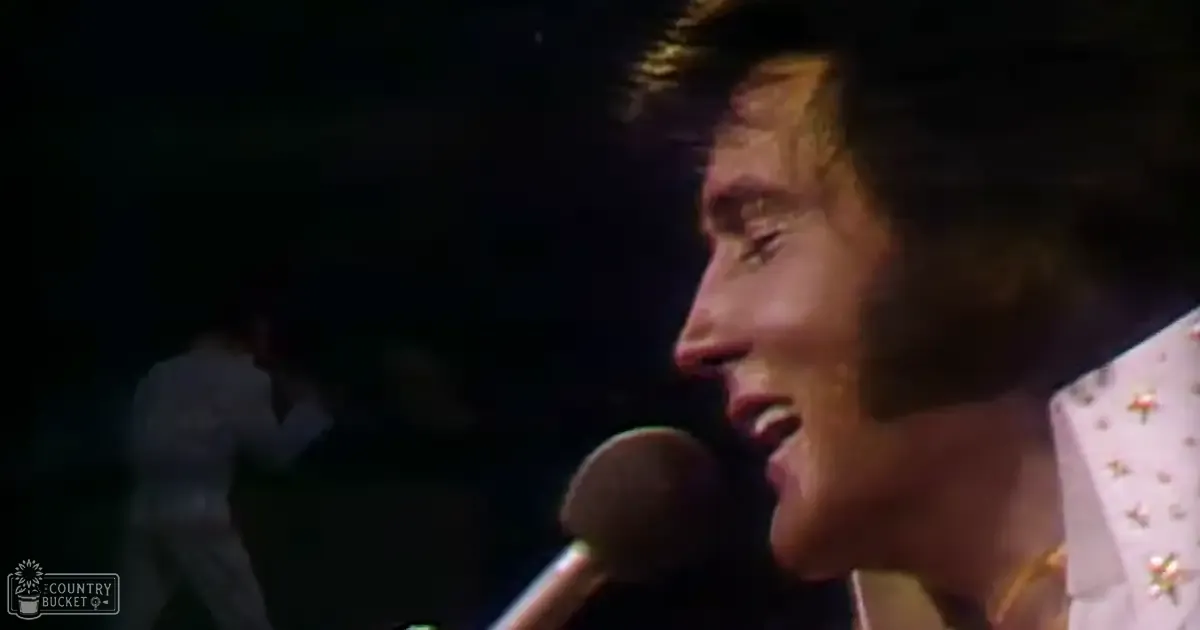 Adult male singer wearing a white 1970s-style shirt with gold stars, performing into a microphone on stage under dim lighting.