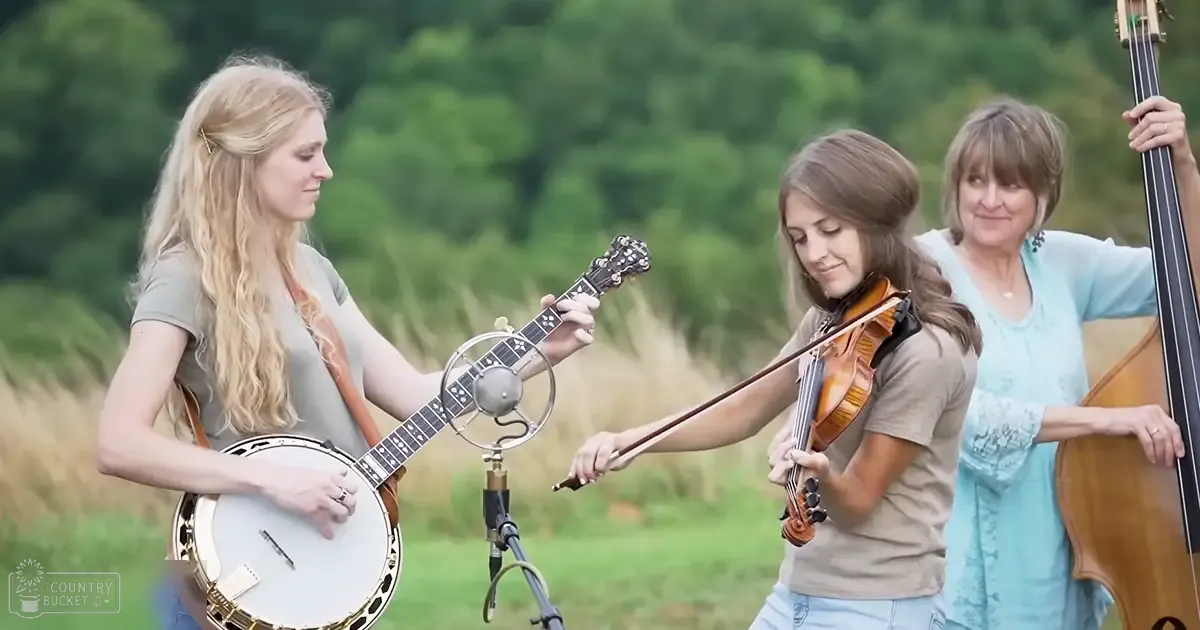 An outdoor musical performance with three women playing banjo, violin, and double bass in a rural setting during daytime.