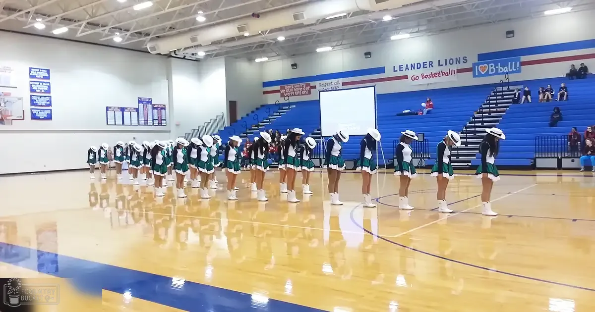 Teenage cheerleaders in a gymnasium at Leander Lions, wearing white hats with black bands and green skirts, standing indoors under artificial lighting during the day.