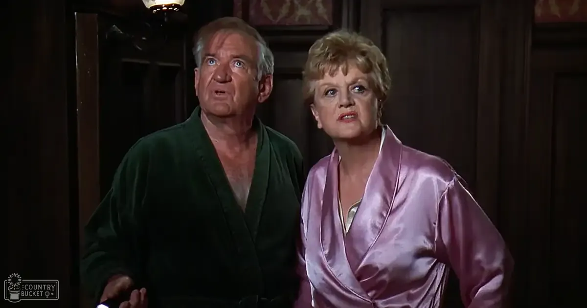 Middle-aged couple in a domestic setting, wearing silk robes; dark green robe with V-neck, pink wrap-style design; wooden door and paneling background.
