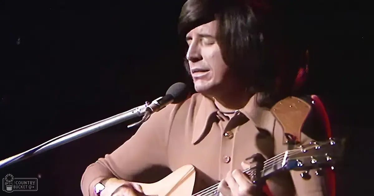 A male adult performer wearing a light brown, collared shirt with a bow tie plays an acoustic guitar on stage under focused lighting.