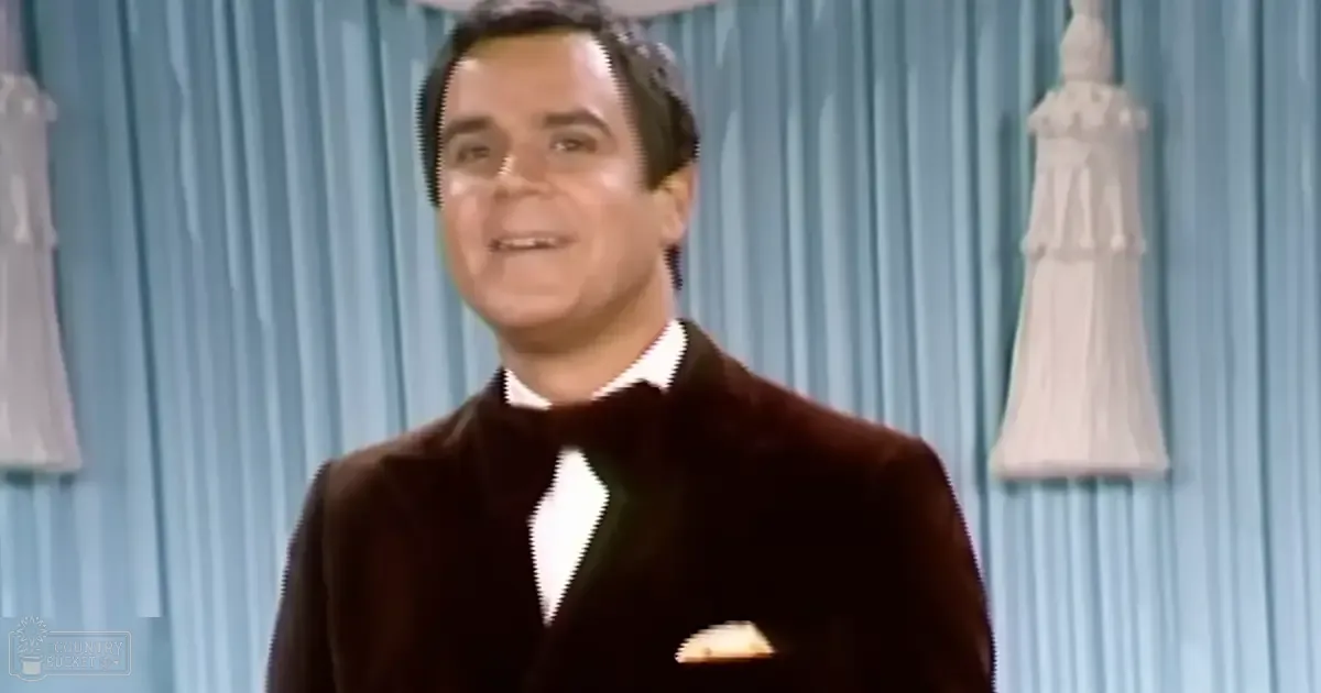 A middle-aged adult male wearing a tailored velvet blazer with a white collared shirt and black bow tie, standing against light blue curtains with decorative elements resembling dresses hanging on stands in an indoor setting.