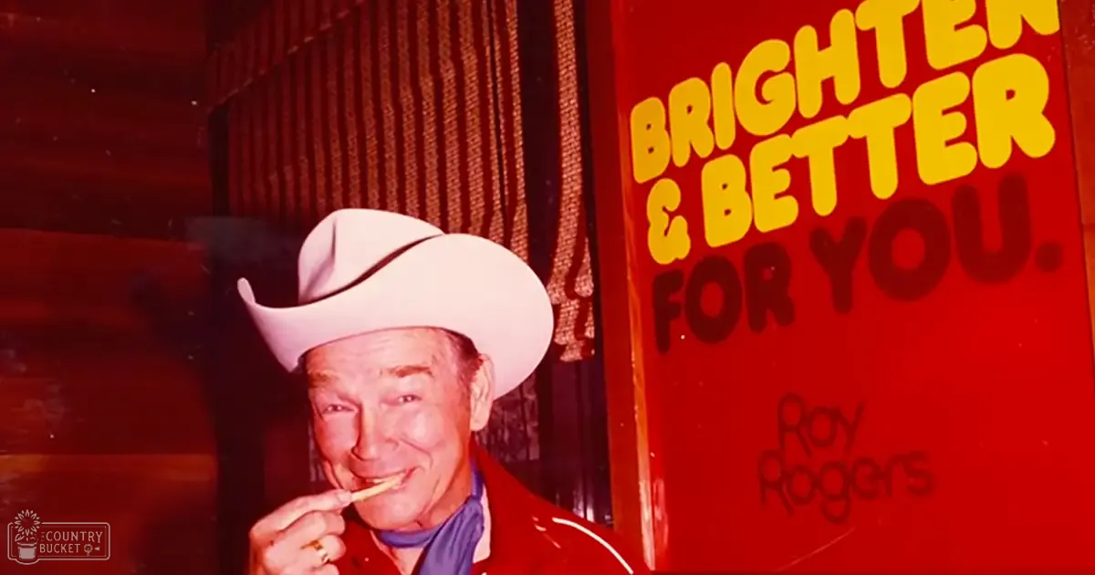 An older adult male in a red mid-20th-century-style outfit, wearing a cowboy hat and holding something near his mouth. The setting appears to be indoors with natural lighting.
