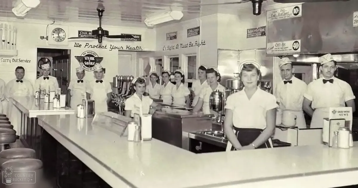 Mid-20th-century diner scene with workers wearing aprons, white shirts, and caps or hats; long white counter, metal condiment shakers, cash register, wall signs reading 'We Protect Your Health' and 'Chili Mac', ceiling fan.