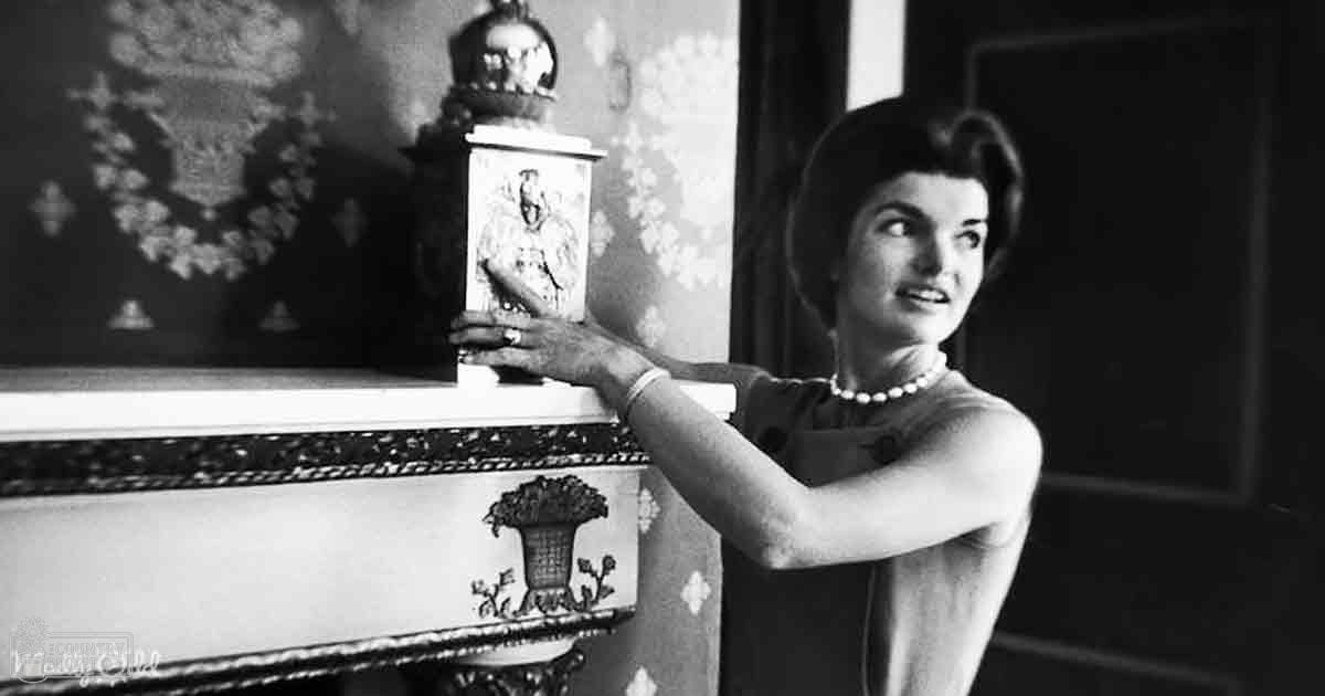 An adult female wearing a sleeveless dress with small circle patterns in mid-20th century style, holding an ornamental box indoors with vintage wallpaper and lighting suggesting daytime